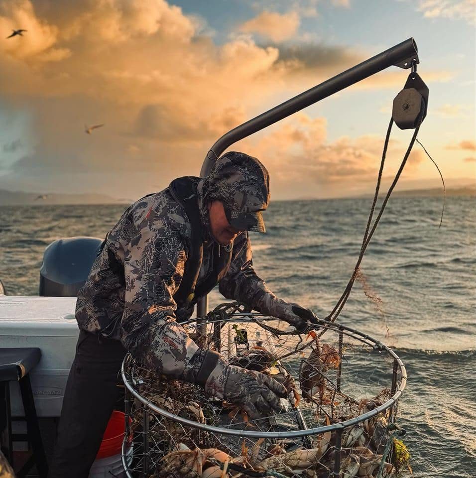 Person in camouflage gear sorting crabs and sea life in a wire trap on a boat at sunset.